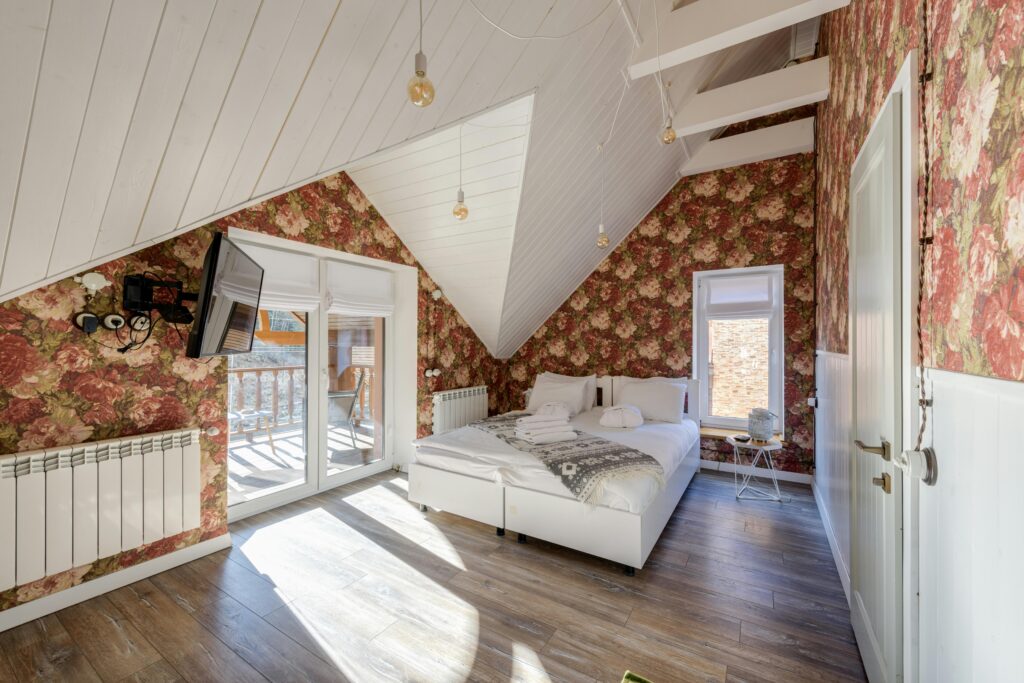 Bright sunlit bedroom with floral wallpaper, white bed, and wooden flooring. Modern and cozy interior design.